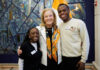 Mercer president seeks input on University’s future Mercer University President Dr. Penny L. Elkins and two students stand smiling together indoors in front of a colorful mural and a flag.