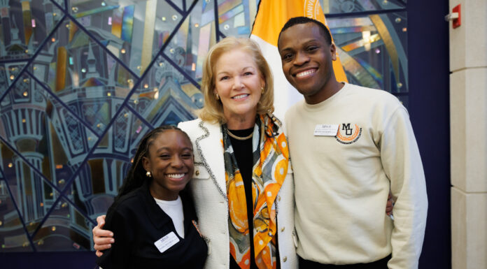 Mercer president seeks input on University’s future Mercer University President Dr. Penny L. Elkins and two students stand smiling together indoors in front of a colorful mural and a flag.