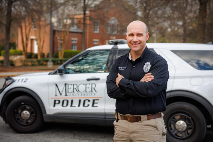 Deputy Chief Jeremy Robinson stands with arms crossed in front of a Mercer University Police vehicle parked on a tree-lined street.