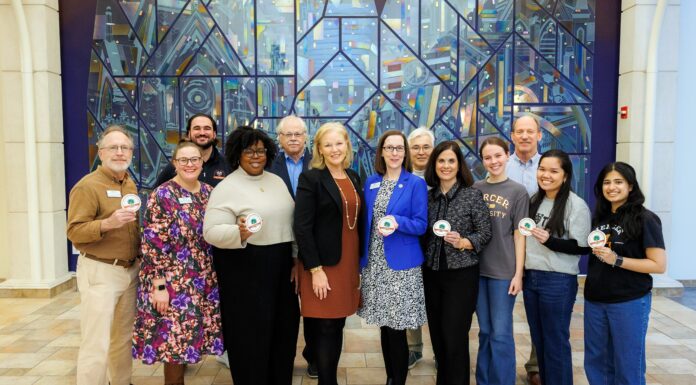 Mercer selected for Carnegie Foundation 2026 Community Engagement Classification Twelve people standing indoors in front of a colorful glass mural, some holding round white badges and smiling at the camera.