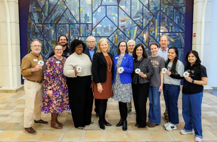 Twelve people standing indoors in front of a colorful glass mural, some holding round white badges and smiling at the camera.