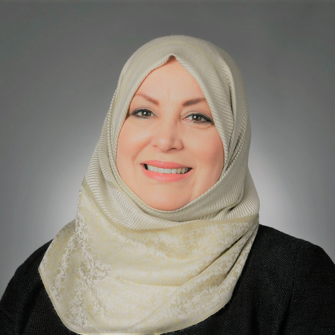 Woman wearing a cream-colored hijab and black top, smiling in front of a plain gray background.
