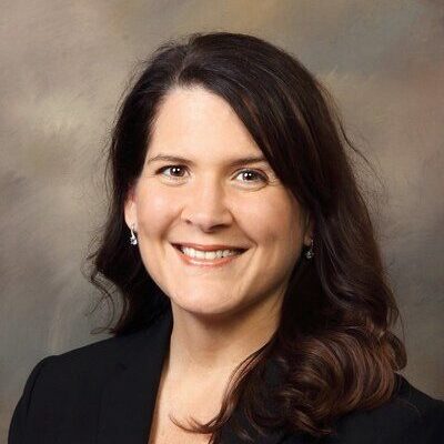 Woman with long brown hair wearing a black blazer, smiling at the camera against a neutral background.