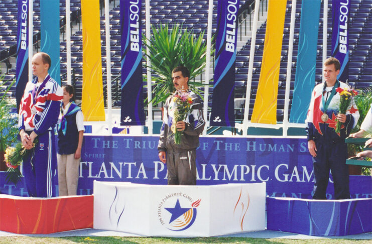 Three male athletes stand on a Paralympic podium with medals and flowers at the Atlanta Paralympic Games.
