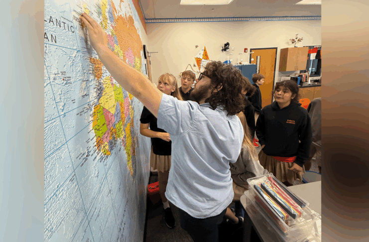 A teacher points to a location on a world map while several students watch attentively in a classroom.