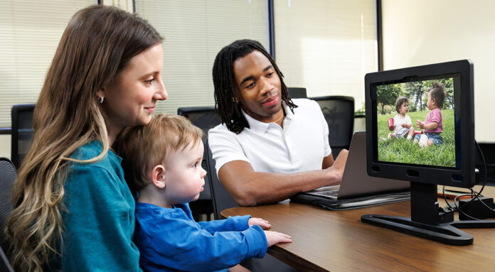A mother holds her child while he watches a video on the EarliPoint device while the technician oversees.