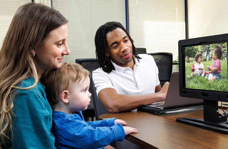 A mother holds her child while he watches a video on the EarliPoint device while the technician oversees.