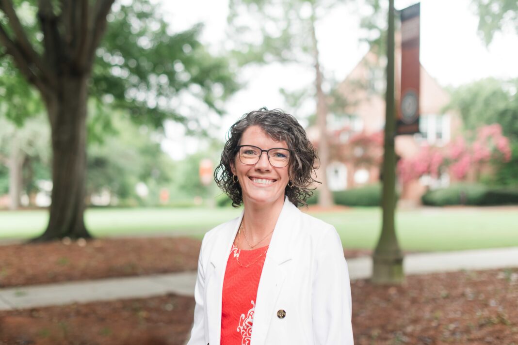 Woman with short curly hair and glasses, wearing a white blazer, standing outdoors on a campus with trees in the background.