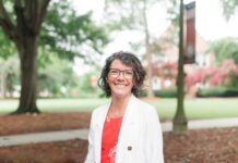 Emmilee Mitchell named interim associate vice president for financial planning Woman with short curly hair and glasses, wearing a white blazer, standing outdoors on a campus with trees in the background.