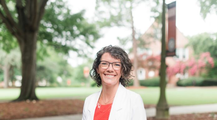 Emmilee Mitchell named interim associate vice president for financial planning Woman with short curly hair and glasses, wearing a white blazer, standing outdoors on a campus with trees in the background.