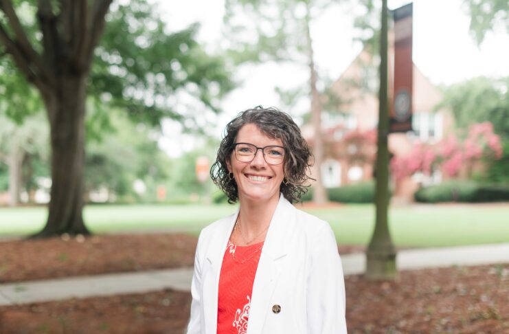 Woman with short curly hair and glasses, wearing a white blazer, standing outdoors on a campus with trees in the background.