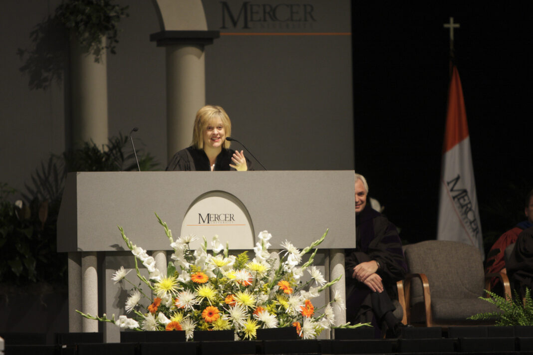 A woman in academic regalia speaks at a podium decorated with flowers at a Mercer University event.