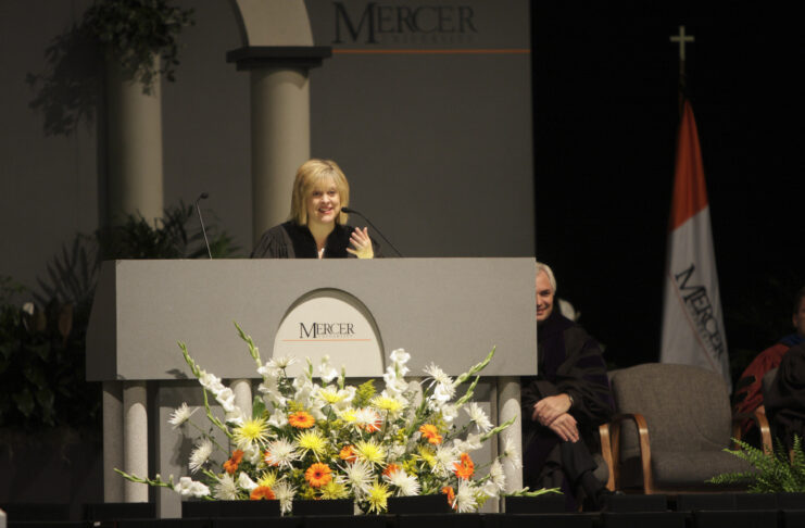 A woman in academic regalia speaks at a podium decorated with flowers at a Mercer University event.