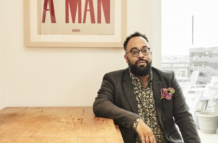 A man with glasses sits at a wooden table beneath a framed sign that reads I AM A MAN in bold red letters.