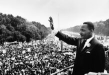 Dr. Joseph M. Hendricks Center for Campus Initiatives to host commemorative program honoring life and legacy of Dr. Martin Luther King Jr. A man in a suit waves to a large crowd gathered outdoors during a historic civil rights demonstration.