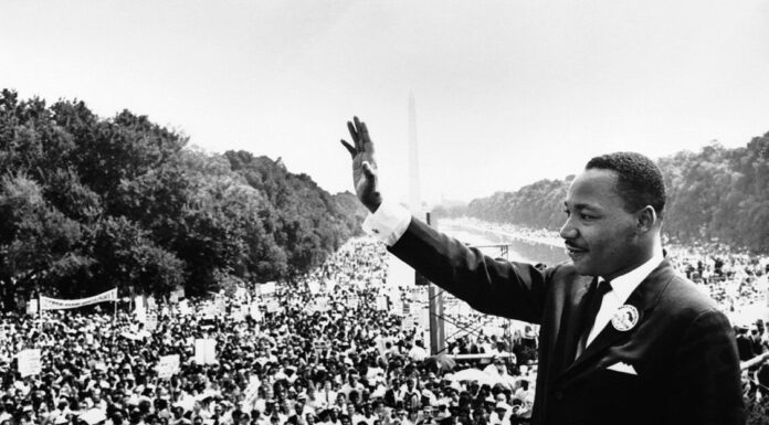 Dr. Joseph M. Hendricks Center for Campus Initiatives to host commemorative program honoring life and legacy of Dr. Martin Luther King Jr. A man in a suit waves to a large crowd gathered outdoors during a historic civil rights demonstration.