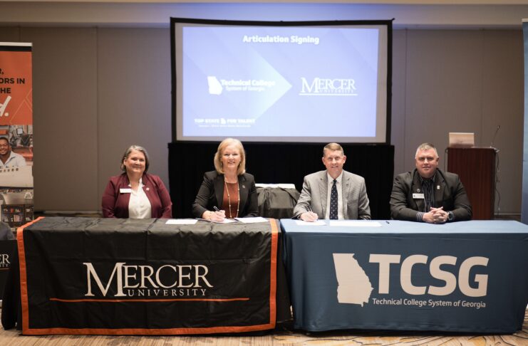 Four people sit at tables with Mercer University and TCSG logos, signing an articulation agreement in a conference room.