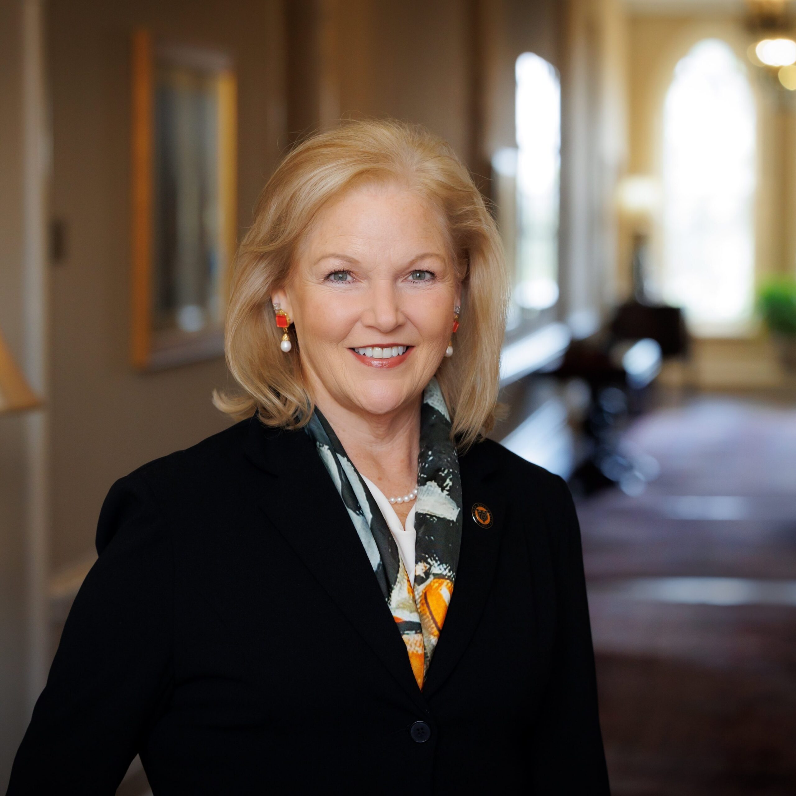 A woman with blonde hair in a black blazer and scarf stands smiling in a well-lit hallway with arched windows.