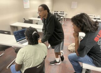 Mercer club provides space for video game creation Three people in a classroom look at code on a laptop, with one person standing and talking while others sit.