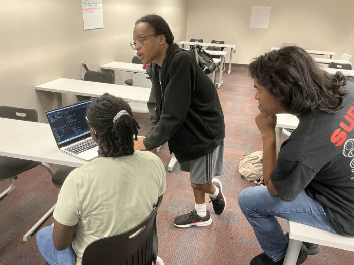 Three people in a classroom look at code on a laptop, with one person standing and talking while others sit.
