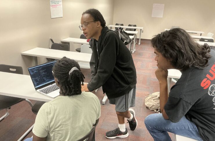 Three people in a classroom look at code on a laptop, with one person standing and talking while others sit.