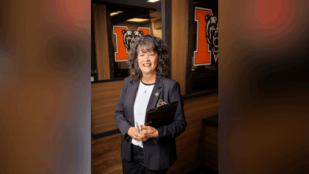 A woman in business attire holds a clipboard, standing in an office with bear logos on glass walls behind her.