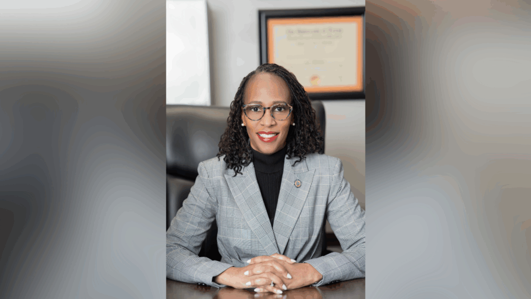 horizontal blurred background (3) A woman in a gray plaid suit sits at a desk, hands folded, with a diploma hanging on the wall behind her.