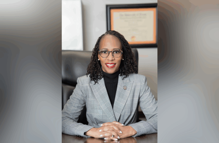 A woman in a gray plaid suit sits at a desk, hands folded, with a diploma hanging on the wall behind her.