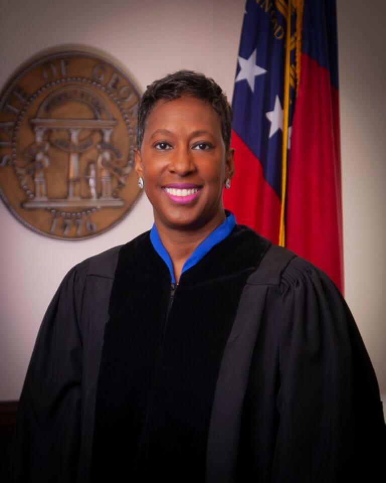 A judge in black robes stands smiling in front of a Georgia state seal and a partially visible Georgia state flag.