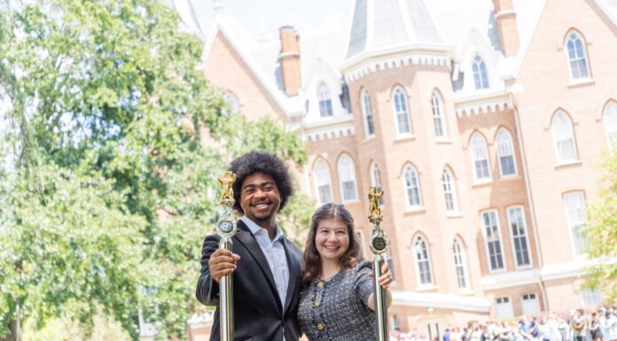 Mercer senior wants to inspire leadership, resilience in others Two people in formal attire holding ceremonial staffs stand outside a large brick academic building, with a crowd behind them.