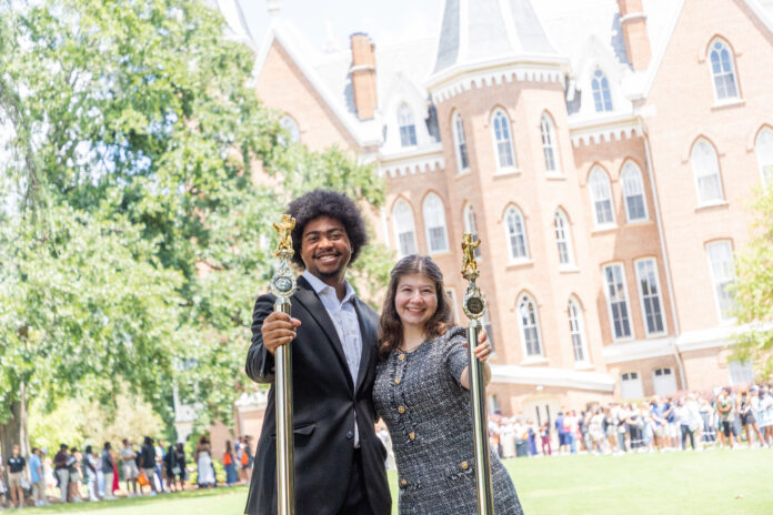 Two people in formal attire holding ceremonial staffs stand outside a large brick academic building, with a crowd behind them.