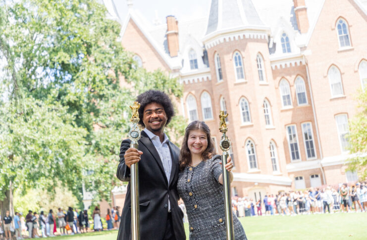 Two people in formal attire holding ceremonial staffs stand outside a large brick academic building, with a crowd behind them.