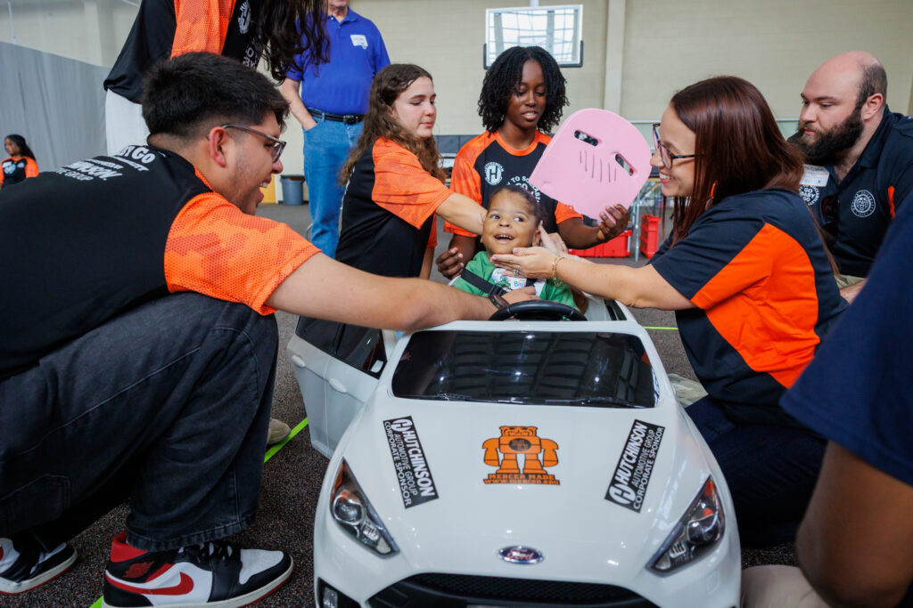 A group of adults assists a smiling child seated in a small white toy car indoors.
