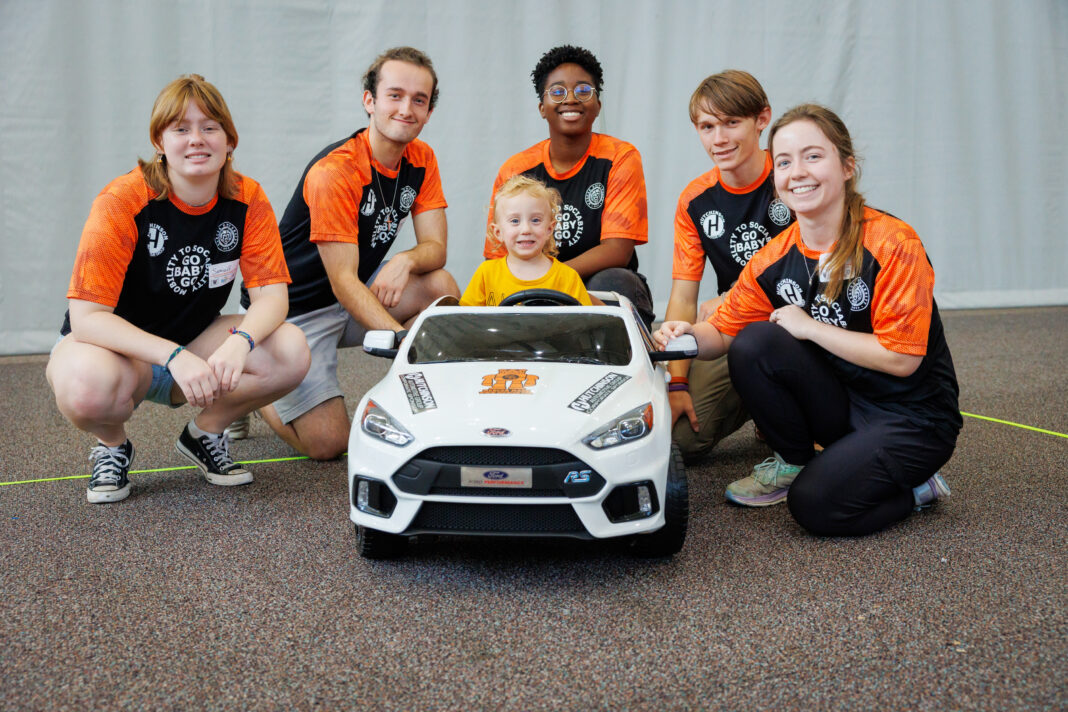 Five young adults and one child pose together behind a small white toy car indoors, all smiling at the camera.