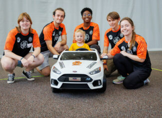 Major milestone as Mercer’s ‘Go Baby Go’ builds 200th toy car for children with limited mobility Five young adults and one child pose together behind a small white toy car indoors, all smiling at the camera.