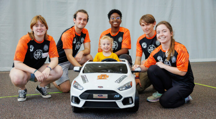 Major milestone as Mercer’s ‘Go Baby Go’ builds 200th toy car for children with limited mobility Five young adults and one child pose together behind a small white toy car indoors, all smiling at the camera.