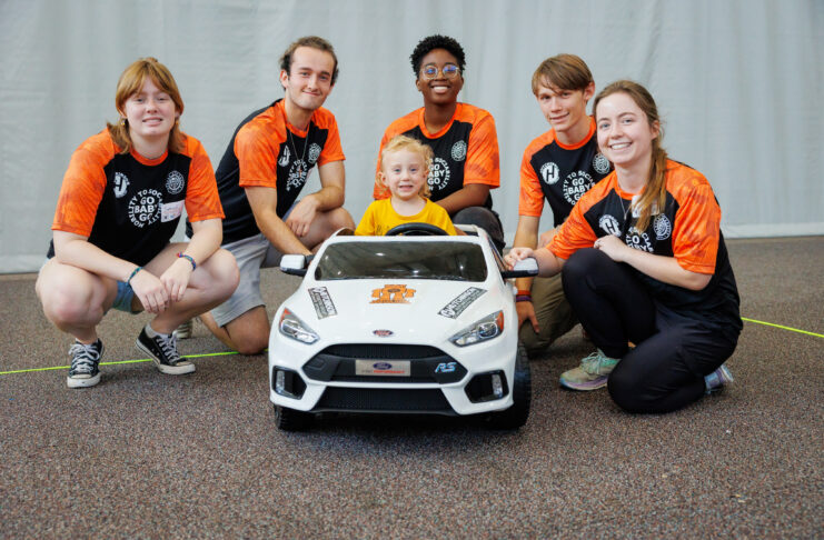Five young adults and one child pose together behind a small white toy car indoors, all smiling at the camera.