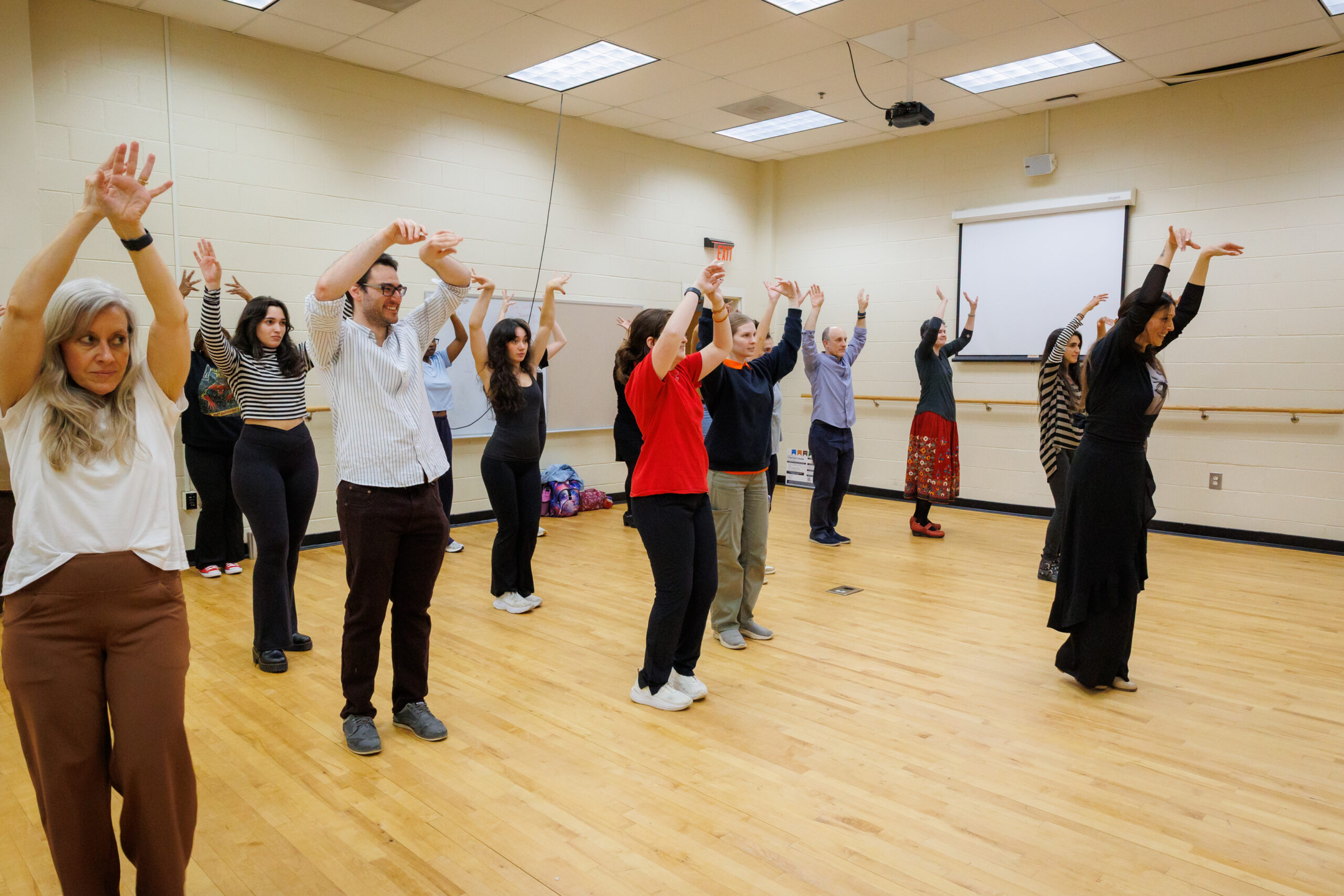 A group of adults stand in a dance studio with arms raised, practicing a synchronized movement.