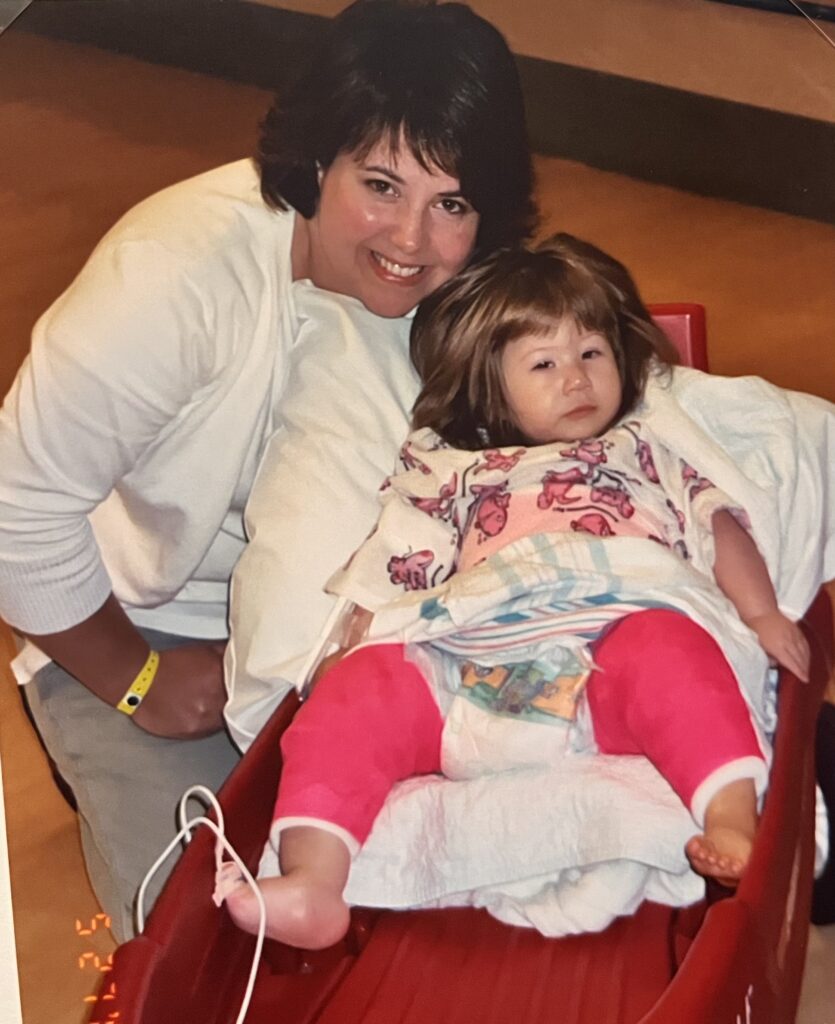 An adult smiles beside a young child resting, with medical supplies and casts on the child's legs.