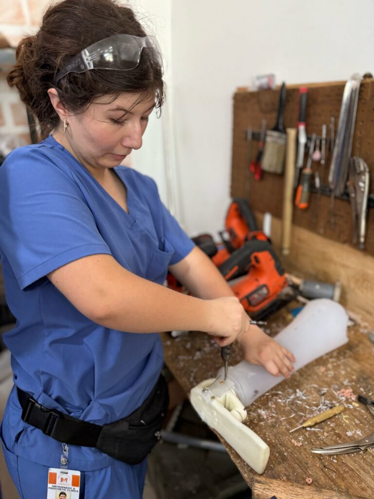 A woman in blue scrubs works on a prosthetic limb at a cluttered workbench with tools and a power drill.