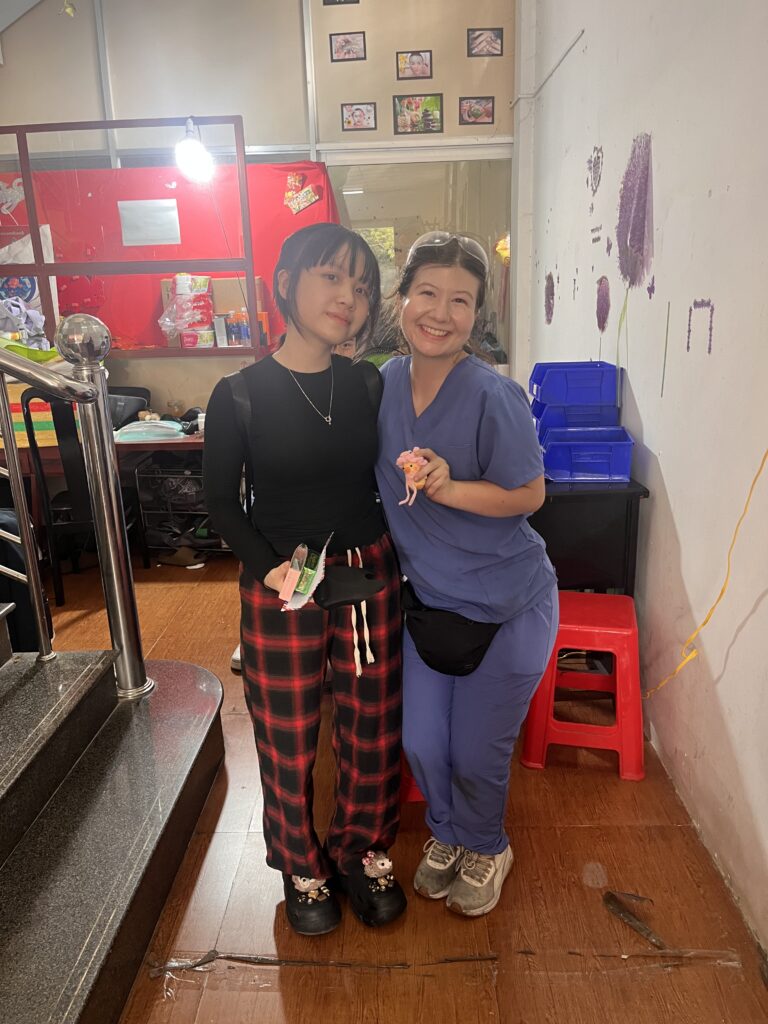 Two women standing indoors, one in casual clothes and the other in scrubs, smiling at the camera.