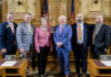 School of Medicine receives over $600,000 to continue fighting opioid epidemic Six people in business attire stand together in a government chamber with flags and wooden desks visible in the background.