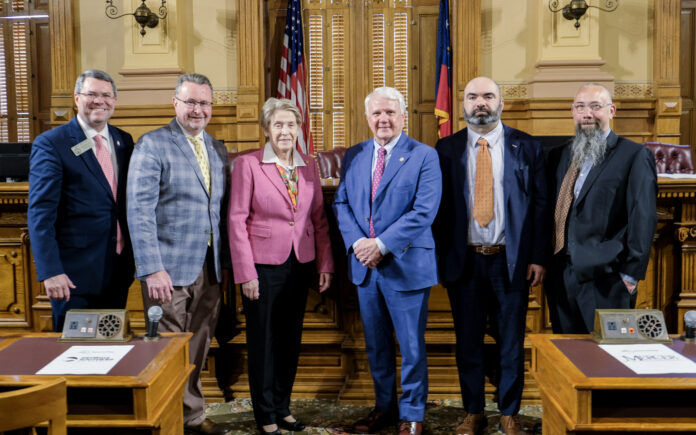 Six people in business attire stand together in a government chamber with flags and wooden desks visible in the background.