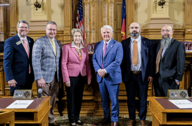Six people in business attire stand together in a government chamber with flags and wooden desks visible in the background.