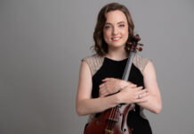 McDuffie Center for Strings welcomes alumna Emily Brandenburg as new director Woman in a black dress holding a violin and smiling, standing in front of a plain gray background.