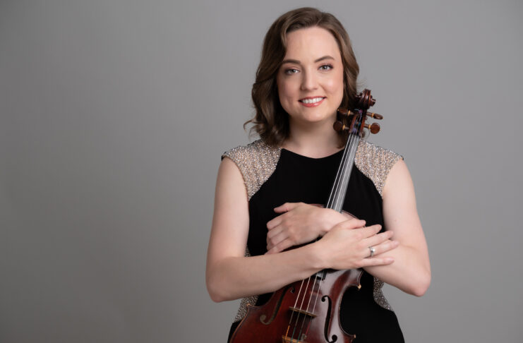 Woman in a black dress holding a violin and smiling, standing in front of a plain gray background.