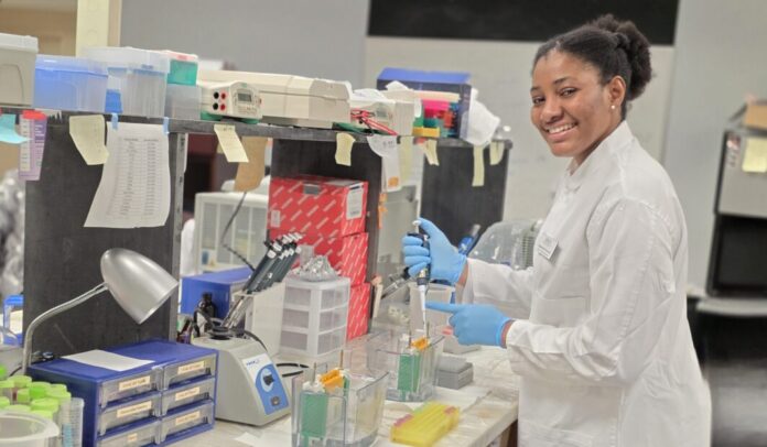 Emmanuella Emmanuella Ezewudo in a lab coat uses a pipette at a laboratory bench with various scientific equipment and supplies.