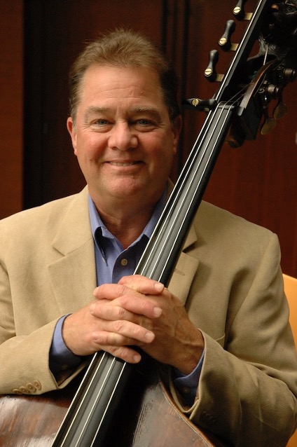 A man in a beige blazer smiles while holding an upright bass in front of a wooden background.