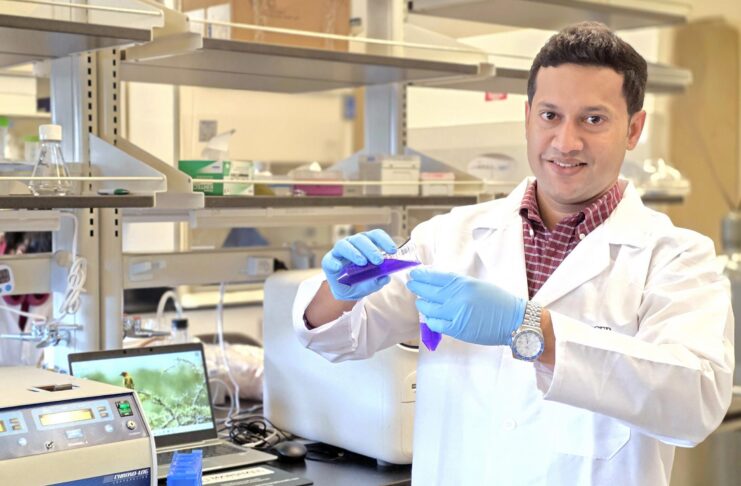 Dr. Raquibul Hasan in a lab coat and gloves handles a test tube with blue liquid in a laboratory setting.