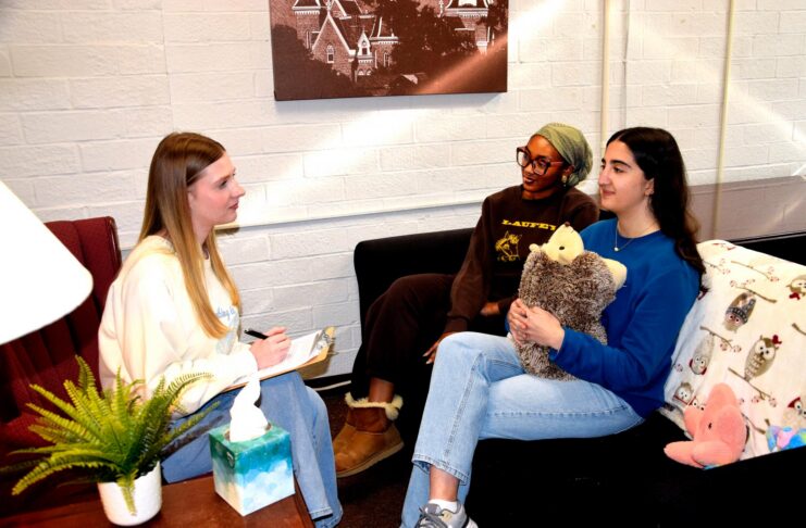 Three women sit in a cozy room talking; one takes notes, while another holds a stuffed animal on the couch.